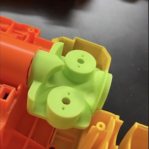 May include: A disassembled toy gun with bright orange, yellow, and green plastic components. The green piece has three circular protrusions with holes. The orange and yellow parts are connected, showing the internal structure of the toy.