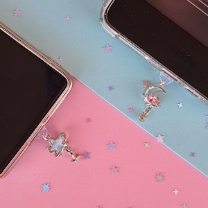 Peut inclure: Deux smartphones et une liseuse Kindle avec des breloques décoratives. L'un des charms présente un croissant de lune et une gemme rose, tandis que l'autre a une étoile et un design en forme de verre. Le fond est rose et bleu avec des confettis en forme d'étoiles.
