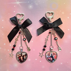 Puede incluir: Dos llaveros en forma de corazón con foto, cadena plateada, dijes de estrella y guitarra, y lazo negro. Los llaveros tienen un cierre plateado y están decorados con cuentas rojas y negras. El fondo es un degradado de rosa y blanco.