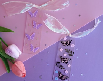 May include: Two clear acrylic bookmarks with butterfly and floral designs. One bookmark features purple butterflies and stars, while the other has black butterflies, leaves, and flowers. Both have iridescent ribbons attached, set against a pink and purple background.