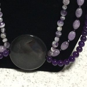 May include: A three-strand necklace and matching earrings set. The necklace features a large, dark gray circular pendant and is composed of various shades of purple and white beads. The earrings have a similar design, with gold-colored hooks.