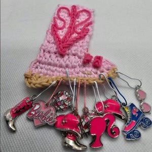 May include: A pink crocheted boot with a heart design and several charms. Charms include a cowboy boot, a heart with "Barbie" text, a disco ball, a hat, a silhouette, and a roller skate. The charms are pink, silver, and blue.