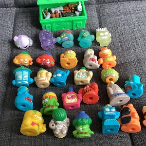 Pode incluir: Uma coleção de pequenas figuras de brinquedo de plástico coloridas, com várias formas e designs, incluindo monstros e criaturas. Uma caixa de plástico verde está aberta, revelando mais brinquedos. Os brinquedos estão dispostos numa superfície cinzenta.