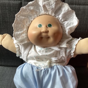 May include: A vintage Cabbage Patch Kid doll with a round face, green eyes, and a white bonnet. The doll is wearing a white top with blue floral accents and a light blue skirt. The doll's arms and hands are a tan colour.