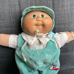 May include: A Cabbage Patch Kids doll with a round face, green eyes, and rosy cheeks. The doll wears a light green corduroy cap, a white collared shirt, and matching light green corduroy overalls featuring a small embroidered sailboat patch.