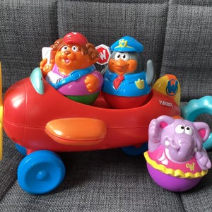 以下が含まれることがあります： 黄色いプロペラ、青い車輪、オレンジ色のアクセントが付いた赤いおもちゃの飛行機。2人の漫画のキャラクターがコックピットに座っています。紫色の象のおもちゃと青いリングも存在します。おもちゃは灰色の表面にあります。