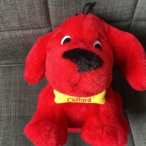 Puede incluir: Un juguete de peluche rojo Clifford, el gran perro rojo, con un collar amarillo que dice "Clifford". El juguete está sentado en un sofá gris.