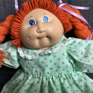 Peut inclure: Une poupée avec des cheveux en laine orange vif coiffés en deux tresses attachées par des rubans violet clair. La poupée a un visage rond, des yeux bleus et des fossettes, portant une robe vert clair à petits motifs floraux et dentelle blanche.