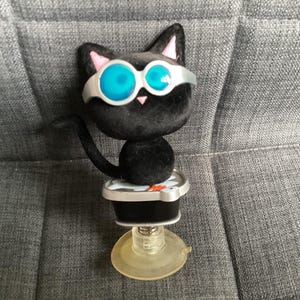 May include: A black cat ornament wearing blue-lensed goggles and pink inner ears. The cat is mounted on a black and silver base with a clear suction cup. The cat's tail is curved, set against a grey background.