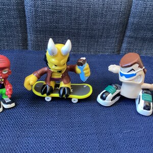 May include: Three toy figures on a blue surface. The figure on the left is red with green clothing and a skateboard. The figure in the middle is yellow with brown clothing and a skateboard. The figure on the right is brown with blue sunglasses and a skateboard.