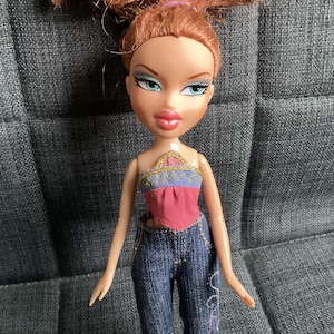 May include: A Bratz doll with auburn hair styled in a high ponytail. The doll wears a pink and purple top with a gold trim and blue denim jeans with embroidered details. The doll has green eyes and full lips.