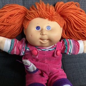 May include: A doll with bright orange yarn hair styled in two pigtails, blue eyes, and a smiling expression. The doll wears a pink corduroy overall outfit over a long-sleeved shirt with pink, white, and teal stripes, featuring a small white animal accent.