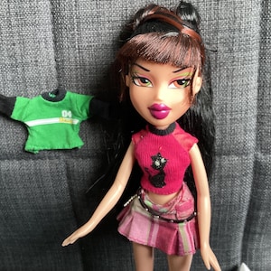 Puede incluir: Muñeca Bratz con pelo largo negro, que lleva una camiseta rosa con un dibujo de gato negro, una falda a cuadros rosa y blanca y una camisa verde y negra. La muñeca tiene maquillaje brillante y posa sobre una superficie gris.