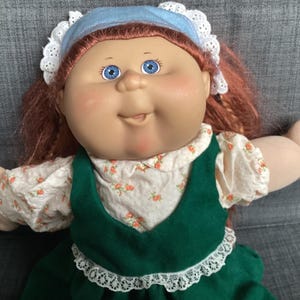 Peut inclure: Une poupée aux cheveux roux, aux yeux bleus et au visage rond. La poupée porte un bonnet bleu clair avec une bordure en dentelle blanche, un chemisier crème à petites fleurs orange et une robe vert foncé avec une bordure en dentelle blanche.