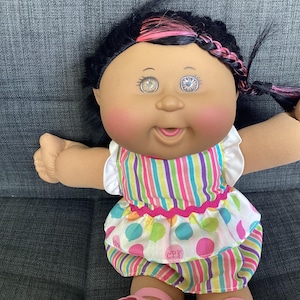 May include: A Cabbage Patch Kids doll with dark hair featuring pink highlights in a braid. The doll has a round face, rosy cheeks, and light-coloured eyes. It wears a dress with a vertical striped bodice and a skirt featuring colourful polka dots.