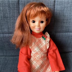 Puede incluir: Una muñeca vintage con cabello rojo y una expresión sonriente. La muñeca viste una camiseta roja de manga larga y un delantal a cuadros con un pequeño acento de acebo verde y rojo. La muñeca está sentada en una silla tapizada gris.