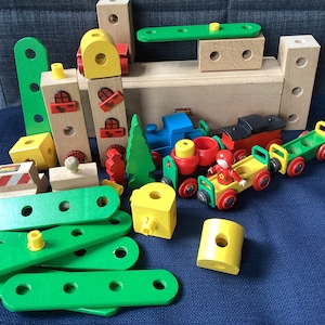 Op de afbeelding: Een set houten speelgoedtreinsporen en treinwagons, waaronder een rode wagon met een geel dak, een blauwe wagon en een groene wagon. De set bevat ook een groene houten brug, een gele houten blok en een rood houten figuur.