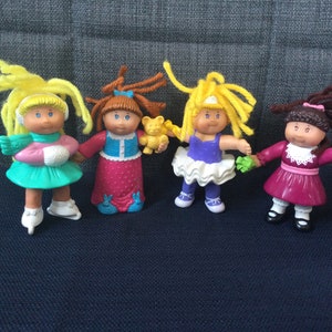 Peut inclure: Quatre poupées Cabbage Patch Kids avec des cheveux en laine. Les poupées portent des tenues différentes, dont une robe verte avec des patins à glace, une robe rose avec une veste bleue, une robe violette avec un tutu blanc et une robe rouge avec un col blanc.