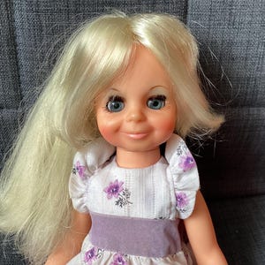 Puede incluir: Una muñeca vintage con pelo largo y rubio y ojos azules. La muñeca lleva un vestido blanco con un estampado floral morado y una cinturilla morada a juego. La muñeca está sentada en una silla tapizada gris.