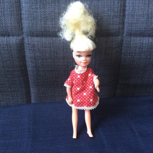 May include: A vintage doll with blonde hair styled in a high ponytail, wearing a red dress with white heart print and white lace trim.