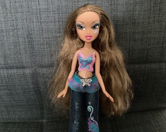 May include: A Bratz doll with long, wavy brown hair and a pink and blue top. She wears blue jeans with a decorative design and a butterfly belt buckle. A small, sparkly diamond is on her cheek.