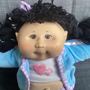 Peut inclure: Poupée Cabbage Patch Kids avec des cheveux noirs en fil de laine tressés en couettes. La poupée a les yeux bruns et les joues roses. Elle porte une veste bleu clair avec une bordure rose et un t-shirt blanc avec un cœur rose.
