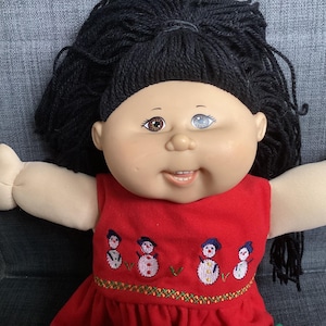 Peut inclure: Une poupée aux cheveux en laine noire, au teint clair et à l'hétérochromie, avec un œil marron et un œil bleu. La poupée porte une robe rouge sans manches brodée d'un motif de quatre petits bonhommes de neige et d'une bordure verte et jaune.