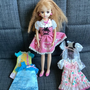May include: A doll with blonde hair and a pink dress with a white collar and black detailing. Several doll dresses are visible, including a blue and yellow dress. The dresses are in various styles.