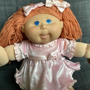 May include: A doll with orange yarn hair styled in two pigtails, each adorned with a light pink satin bow. The doll has bright blue eyes, a small nose, and a smiling mouth. It wears a light pink velvet dress with ruffled sleeves and a bow.