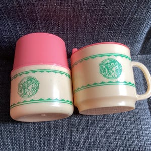 May include: A vintage baby bottle and mug set. The bottle has a pink lid, and both items are cream-coloured with a green border and emblem. A classic design for feeding.