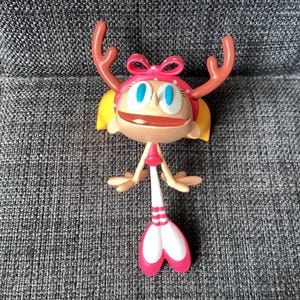 以下が含まれることがあります： ピンクのリボン、角、ピンクと白のストライプの尾が付いた、カラフルな漫画のフィギュア。 このフィギュアは大きな青い目、金色の髪、笑顔の表情をしています。 このフィギュアは灰色の表面に立っています。