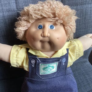 Peut inclure: Poupée Cabbage Patch Kids avec une peau claire, des yeux bleus et des cheveux blonds bouclés. La poupée porte une chemise jaune à carreaux vichy sous une salopette en jean bleu. La salopette porte une étiquette verte et blanche avec l'inscription "Cabbage Patch Kids."