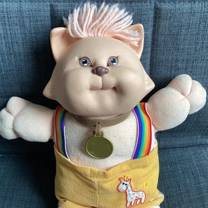Puede incluir: Muñeca de gato de peluche con cara melocotón claro, pelo rosa y ojos azules. Viste un atuendo de pana amarilla con tirantes arcoíris y un medallón dorado. Un caballo de dibujos animados blanco y naranja está en el atuendo.