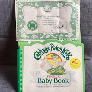 May include: A Cabbage Patch Kids sticker baby book with a stork carrying a baby on the cover. The book has a green spine and a certificate of birth is on top. The certificate is green and white.