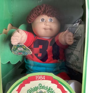 Peut inclure: Une poupée Cabbage Patch Kids de 1984 avec des cheveux en laine brune, portant un haut rouge avec le numéro 31 en noir et un pantalon bleu. La poupée est dans son emballage d'origine en carton vert et blanc, incluant un certificat d'adoption.