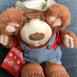May include: A plush bear doll with brown fur, a smooth face, and a moustache. The bear wears a straw cowboy hat, a red shirt, blue and white pinstriped overalls with metal buttons, a red paisley bandana, and brown moulded plastic boots.