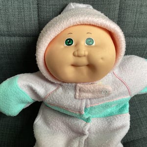 Op de afbeelding: Een vintage Cabbage Patch Kid pop met een lichtroze en witte jumpsuit met capuchon en mintgroene accenten. De pop heeft een rond gezicht, groene ogen en een lichte glimlach. Een klassiek verzamelobject.