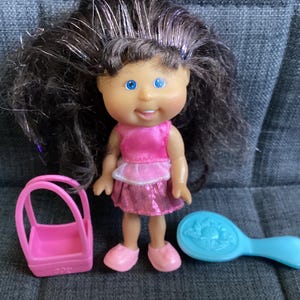 Puede incluir: Una muñeca pequeña con cabello castaño y ojos azules, vestida con un vestido y zapatos rosas. Un bolso rosa y un cepillo azul están al lado de la muñeca. La muñeca tiene una expresión sonriente.