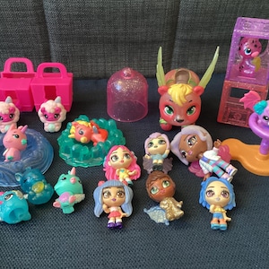 Può includere: Una collezione di piccoli giocattoli di plastica colorati, tra cui un unicorno, un drago, una sirena e una varietà di bambole. I giocattoli sono disposti su un tavolo con uno sfondo rosa e bianco.