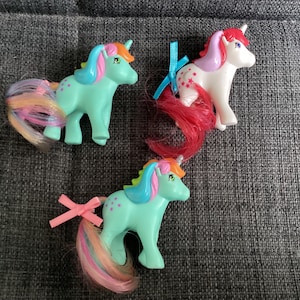 May include: Three plastic unicorn figurines with colourful manes and tails. Two are mint green with orange and blue manes, one with a pink bow. The third is white with a red mane, purple accents, and a blue bow. Each features painted eyes and star or dot symbols.