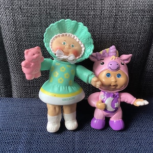 Puede incluir: Dos muñecas Cabbage Patch Kids, una lleva un vestido verde con flores amarillas y sostiene un oso de peluche rosa, la otra lleva un disfraz de unicornio rosa con un diseño de luna y estrella morado y azul.