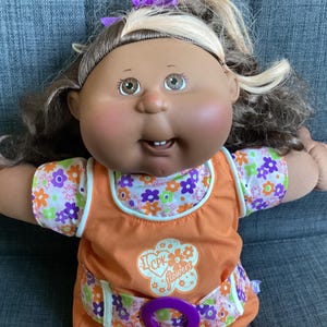 May include: A Cabbage Patch Kids doll with brown hair, light skin, and brown eyes. The doll wears an orange sleeveless dress over a white shirt with a colourful floral pattern. A graphic on the dress reads I CPK flowers with a purple ring at the waist.