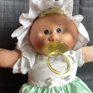 Puede incluir: Una muñeca bebé vintage con una cara suave y redondeada, que lleva un gorro blanco con ribete de encaje y un vestido verde menta. La muñeca tiene ojos marrones y un chupete amarillo. Los brazos de la muñeca están extendidos.
