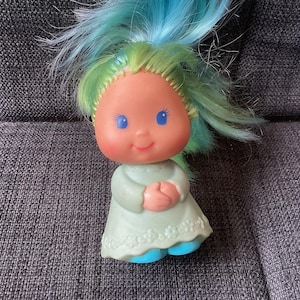 May include: A vintage doll with a smiling face, blue eyes, and blue and green hair. The doll wears a light green dress with a floral hem and blue shoes. The doll's hands are clasped in front of it.