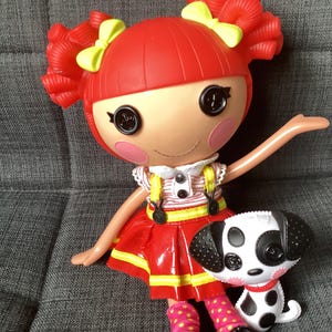 May include: A Lalaloopsy doll with bright red hair styled in pigtails with yellow bows, black button eyes, and pink cheeks. The doll wears a red skirt with yellow trim and a striped top with yellow straps, accompanied by a small black and white dog.
