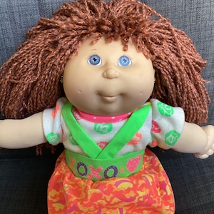 Könnte beinhalten: Eine Puppe mit rundem Gesicht, blauen Augen und strukturiertem rotbraunem Wollhaar. Die Puppe trägt ein weißes Oberteil mit grünen und rosa kreisförmigen Mustern, einen hellgrünen Bund mit dem Text XOXO und einen orangefarbenen Rock mit gelbem Muster.