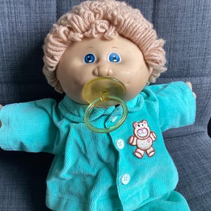 May include: A vintage Cabbage Patch Kids doll with blonde yarn hair, blue eyes, and a dummy. The doll wears a teal corduroy outfit with a teddy bear patch. The doll is sitting on a grey chair.