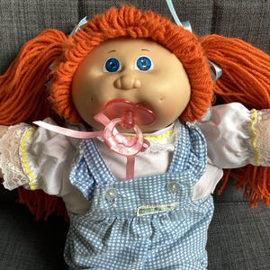 Könnte beinhalten: Eine Vintage-Puppe mit orangefarbenen Zöpfen aus Garn, blauen Augen und einem Schnuller. Die Puppe trägt eine weiße Bluse mit Spitzenbesatz und ein blau-weiß kariertes Latzkleid. Ein rosa Band ist am Schnuller befestigt.