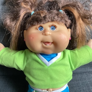 May include: A vintage doll with brown pigtails, blue eyes, and rosy cheeks. The doll is wearing a green sweater with a white and blue trim, and blue trousers. The doll has pink earrings and is smiling.