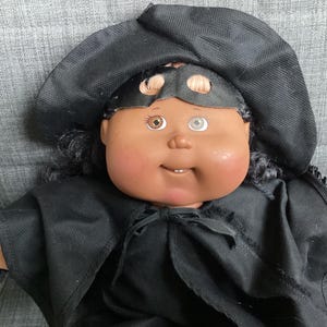 May include: A vintage doll with dark skin, black hair, and a black outfit. The doll wears a black hat and a mask with eye holes. The doll has one brown eye and one blue eye. The doll's outfit includes a cape.